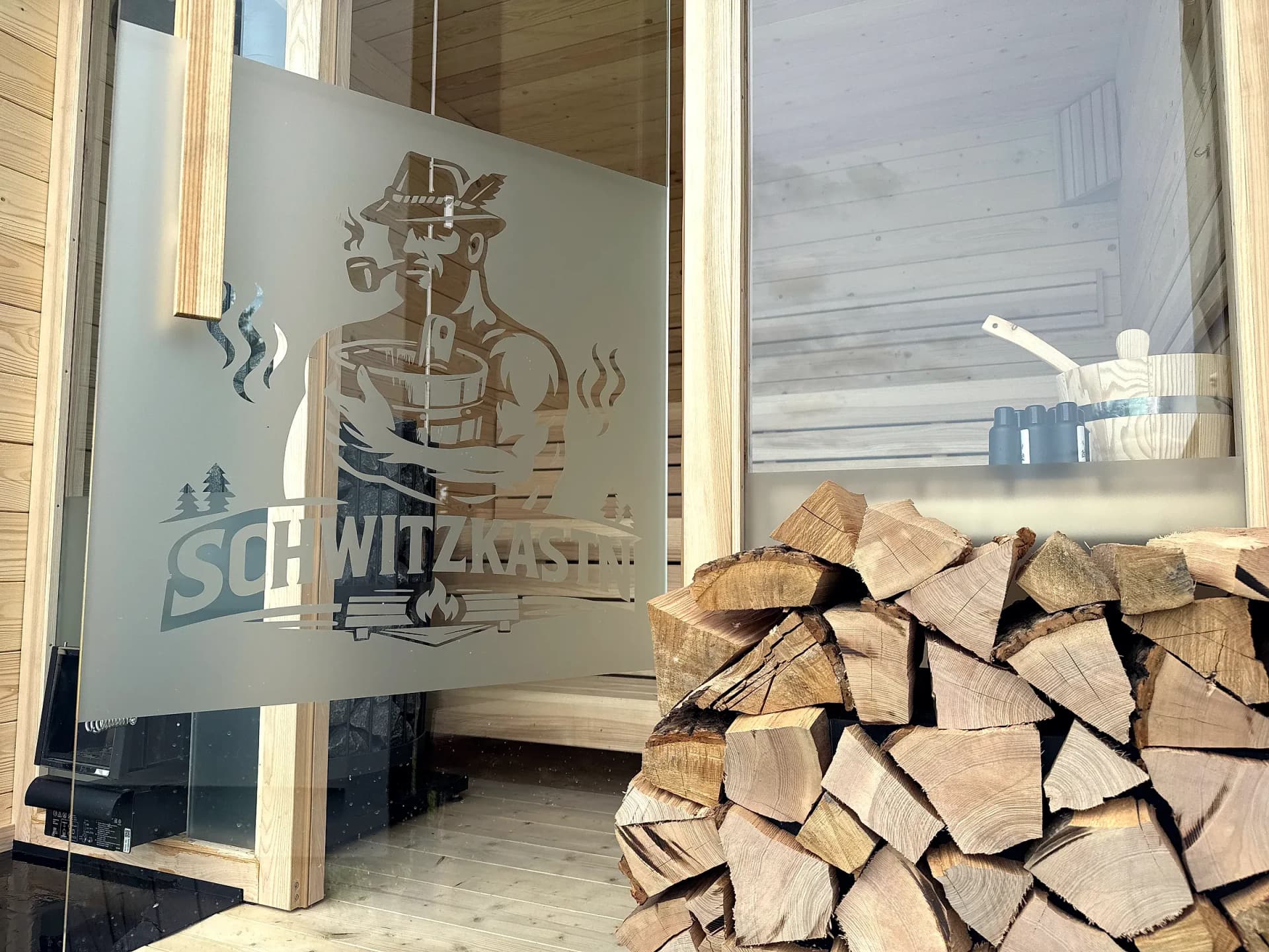 Gestapeltes Brennholz neben der geätzten SCHWITZKASTN-Glaswand