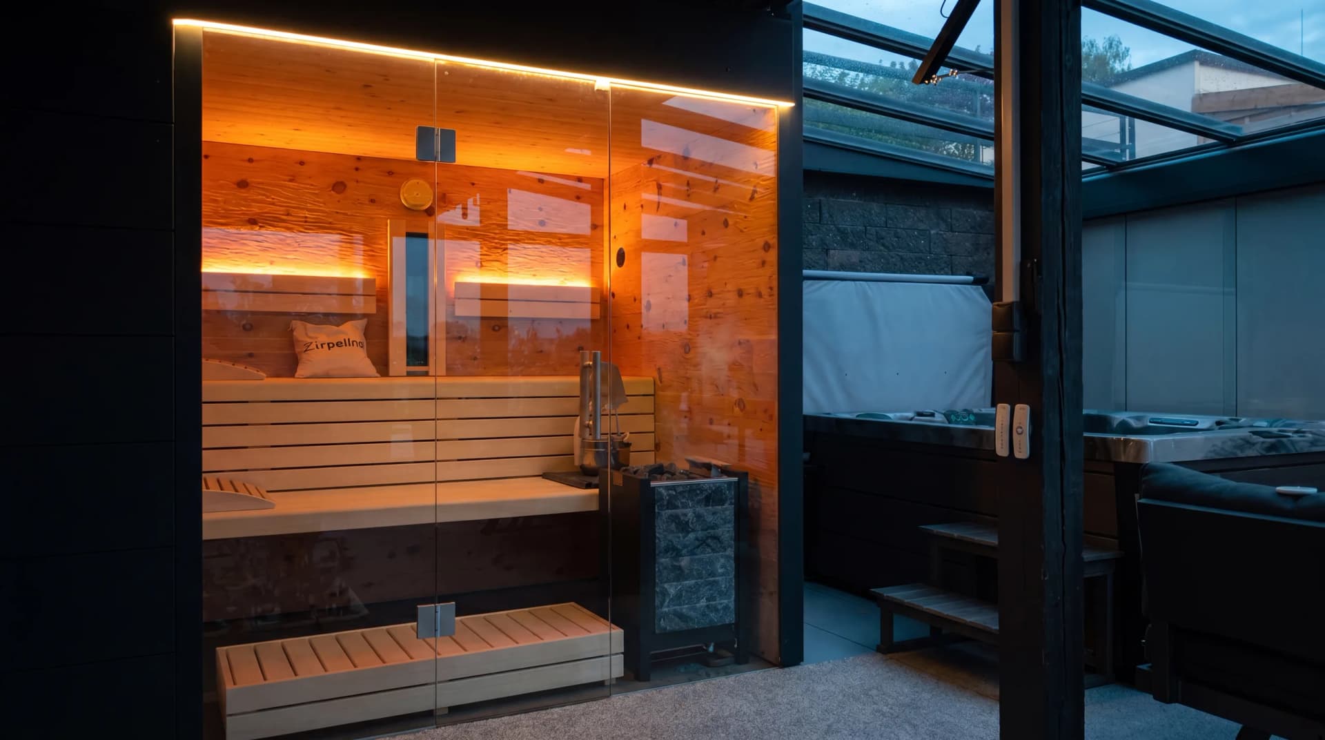 Premium Außensauna auf überdachter Terrasse mit Glasfront, warm beleuchtetem Zirbenholz-Innenraum und Saunaofen in der Abenddämmerung – Gartensauna von Hein Wellnesskonzepte