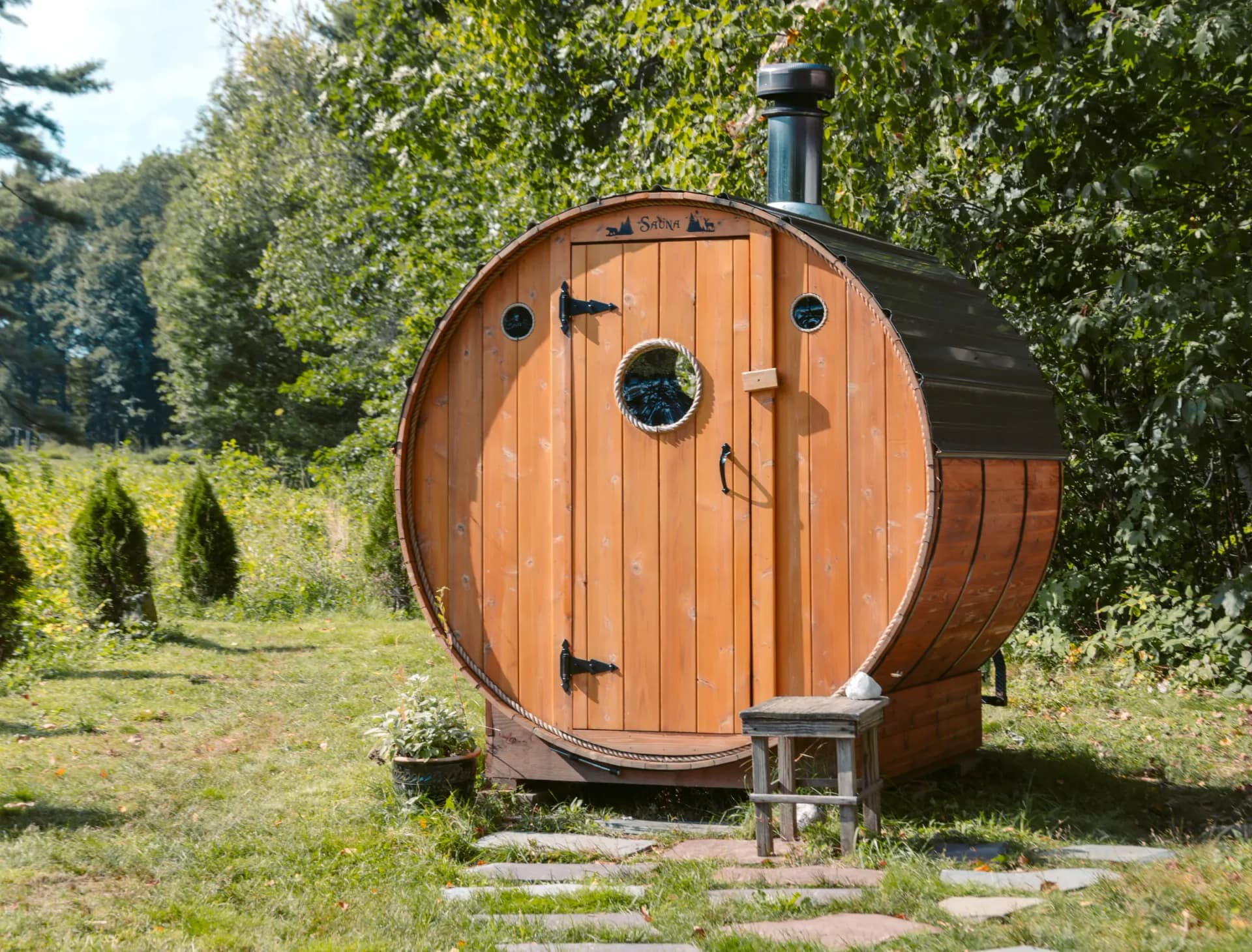 Symbolbild: typische Fasssauna aus Holzdauben mit Metall-Spannbändern, Ofenrohr und runder Stirnseite im Garten. Keine Sauna aus unserer Fertigung