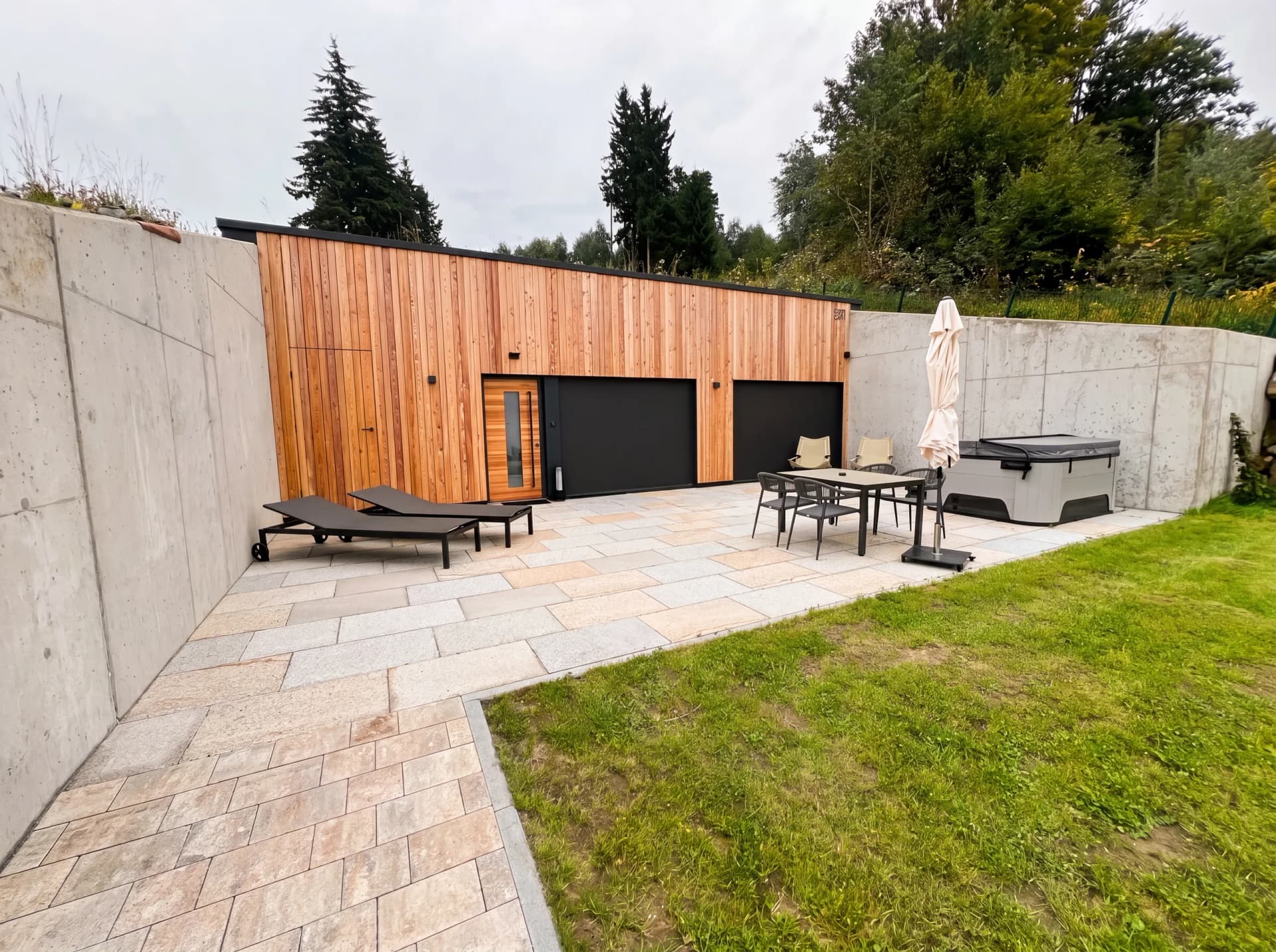 Moderne Gartensauna mit Lärchenholz-Fassade auf gepflasterter Terrasse mit Loungemöbeln und Whirlpool, eingefasst von Sichtbeton-Mauern – Außensauna von Hein Wellnesskonzepte