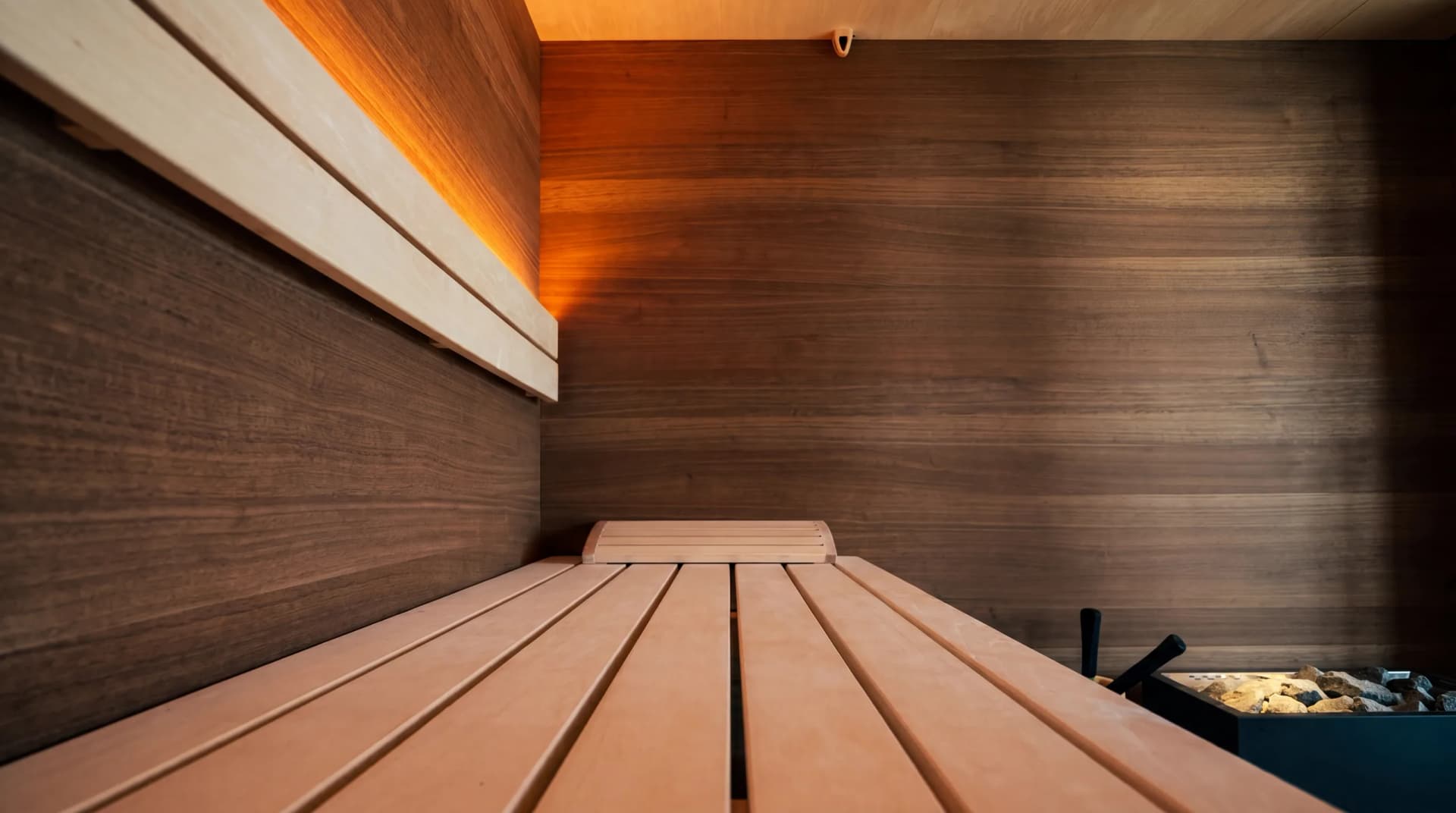 Obere Liegebank einer maßgefertigten Sauna mit ergonomischer Kopfstütze, warmem LED-Lichtband und Nussbaum-Wandpaneelen – Saunainnenausbau von Hein Wellnesskonzepte