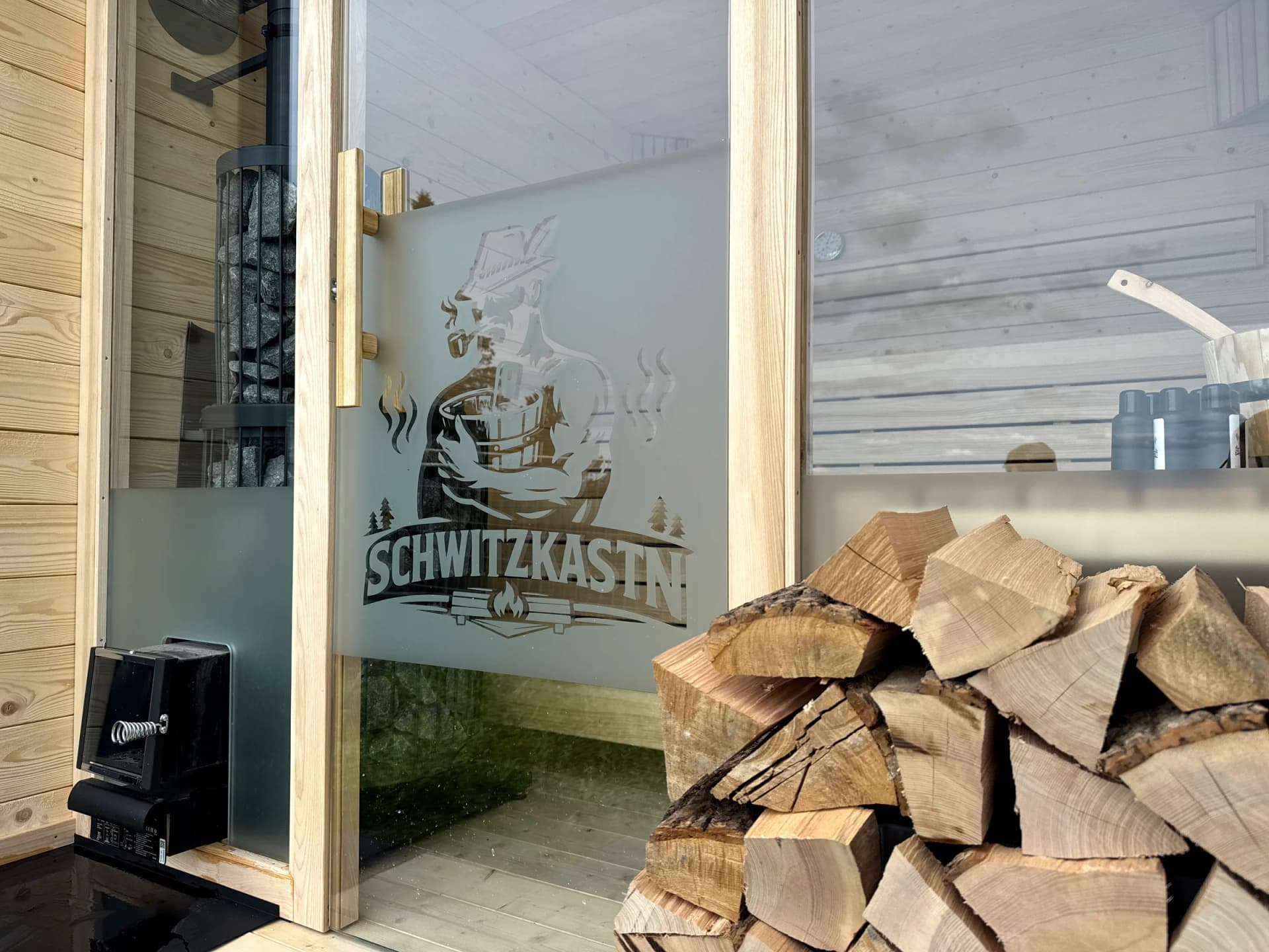 Mattierte Schwitzkastn-Glastür mit Logo, daneben aufgestapeltes Brennholz für den Saunaofen